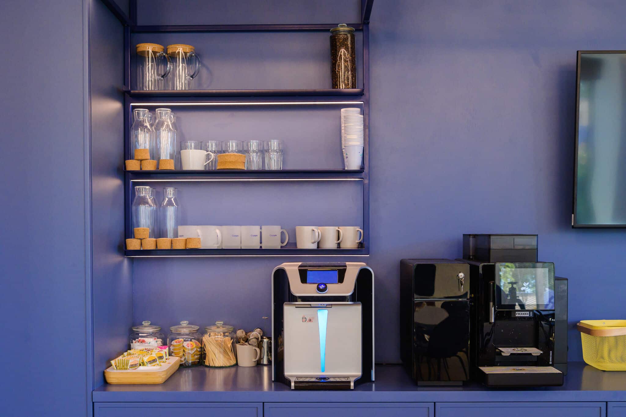 A modern coffee and tea station with glass jars, mugs, water bottles, a water dispenser, a coffee machine, and various supplies on blue shelves and counter.