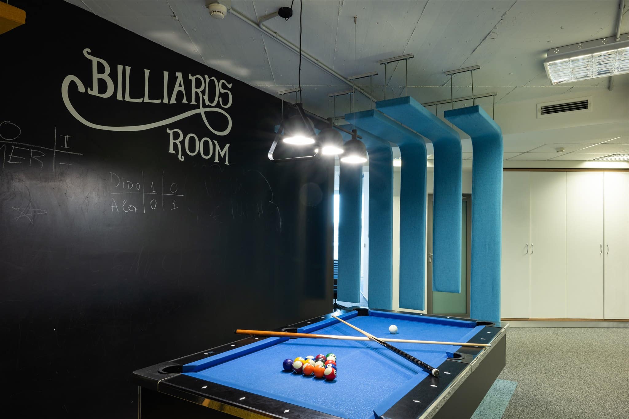 A billiards room with a blue pool table, pool cues, racked balls, a black chalkboard wall labeled "Billiards Room," and blue acoustic panels on the ceiling.