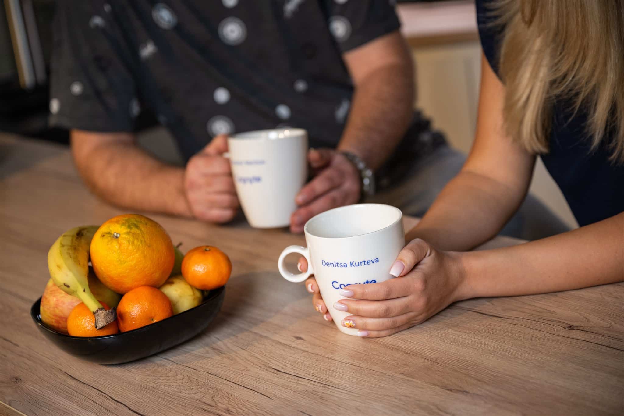 Two people sit at a wooden table holding white mugs, with a black bowl of assorted fruit including bananas, oranges, and apples in front of them.